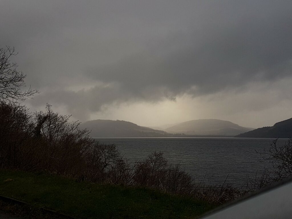 Blick über Loch Ness auf umliegende Berge. Die Wolken sind dunkelgrau und hängen sehr tief, über den Bergen gibt es etwas diffuses Sonnenlicht.