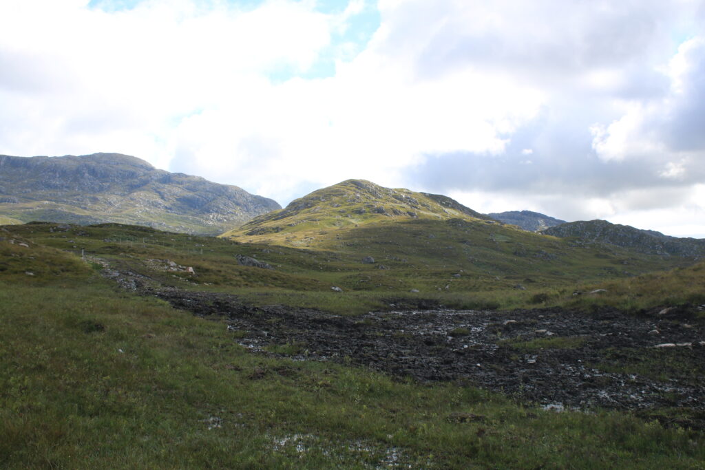 Matschkuhle bei Loch an Nostarie mit Blick in die Highlands. Im Vordergrund ist dunkelbraune, weiche, nasse Erde mit vielen Pfützen aus Wasser. Dazwischen sind einige Grasbüschel. Im Hintergrund sieht man die Hügel, mit grünem Gras bedeckt, außerdem sieht man einige steinigen Stellen und dem Gras. Der Himmel ist blau mit Wolken. 