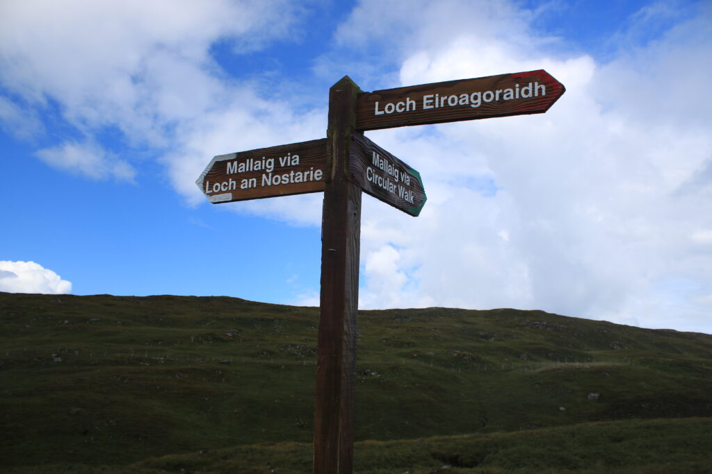 Wegweiser aus Holz am Loch an Nostarie. Es werden 3 Richtungen angezeigt. Im Hintergund sind grüne, mit Gras bewachsene Hügel, blauer Himmel und weiße Wolken