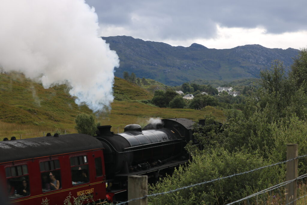 Im Vordergrund sieht man die Lok und einen Wagen vom Jacobite Steam Train.  Die Lok stößt dampf aus, der Wagon ist in dunkelrot angemalt. Im Hintergrund sieht man weiße Häuser und Berge. 