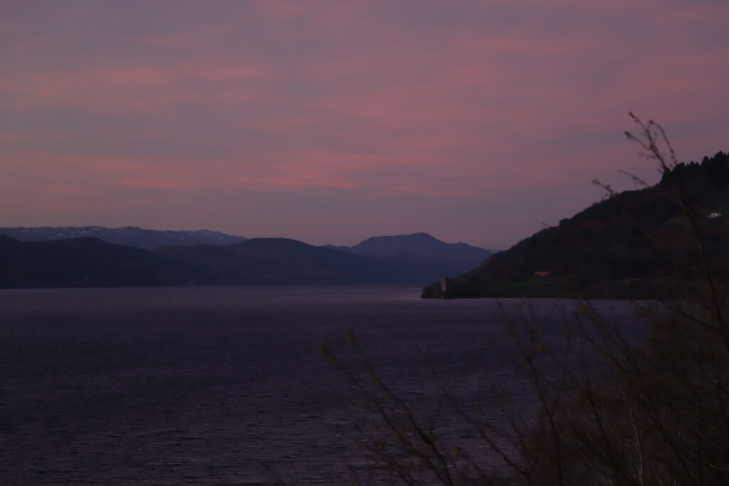 Sonnenaufgang über Loch Ness. Der Himmel ist in Rot und Rosatönen gefärbt. Über dem See steigt leicht Nebel auf. Die Berge sind gräulich und dunkel