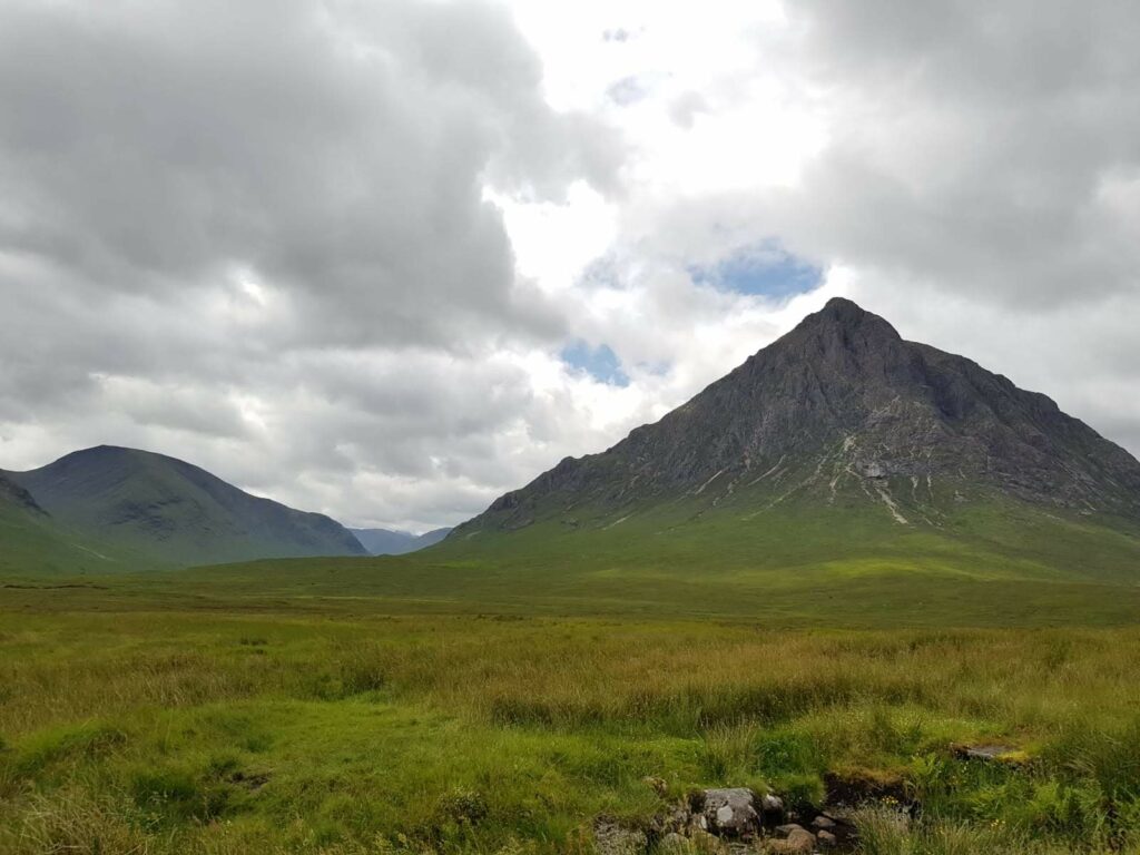 Im Vordergrund ist grüne Graslandschaft, im Hintergrund ragen zerklüftete, dunkelgraue, spitze Berge in den Himmel. Der Himmel ist bewölkt. 