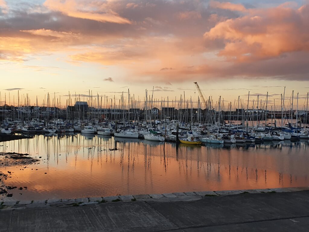 Der Hafen von Howth im Abendlicht. Die Untergehende Sonne und Segelboote spiegeln sich im Wasser. 