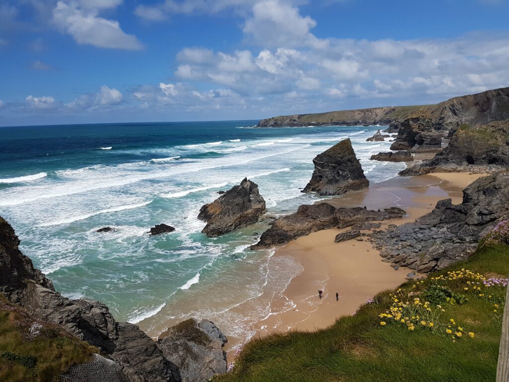 Bedruthan Steps von oben. Das Wasser läuft gerade ab und legt den Strand frei. Der Himmel ist Hellblau und das Wasser Türkis.