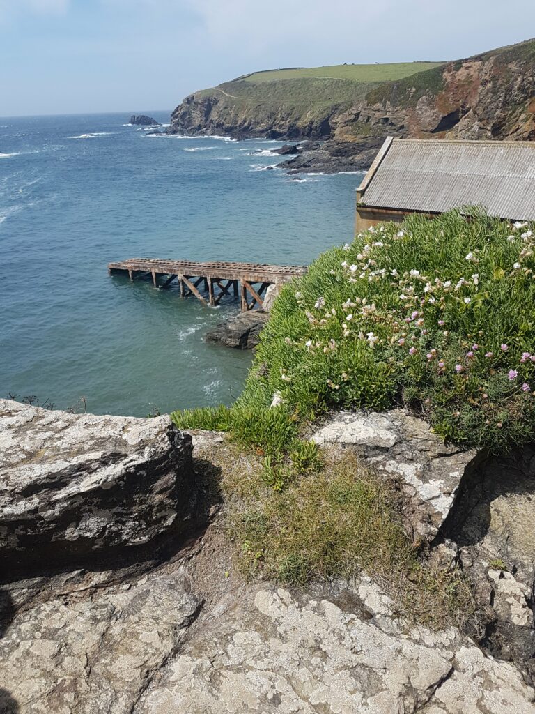 Blick von der felsigen Steilküste auf die Lifeboat Station. Ein langer Steg reicht ins türkise Wasser hinein. 