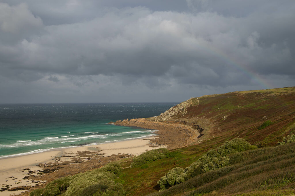 Küstenabschnitt in Cornwall. Die Sonne strahlt das Land an, während über dem Wasser dunkelgraue Regenwolken hängen. Man kann einen leichten Regenbogen auf der rechten Bildseite sehen. Das Wasser ist Türkis und dunkelblau. 