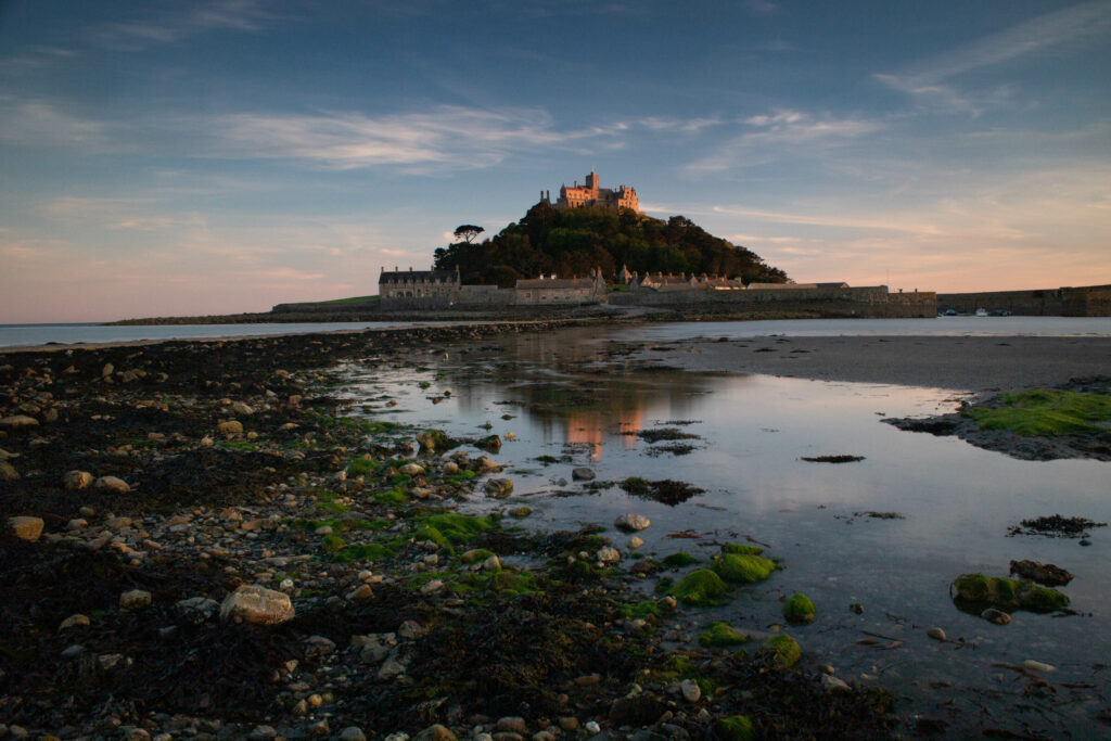 Das Herrenhaus St. Michaels Mount im Abendlicht vom Festland aus fotografiert. Der Himmel ist blau und leicht orange-rosa. Es ist Ebbe, man kann mit grünem Seegras bedeckte Steine sehen. 