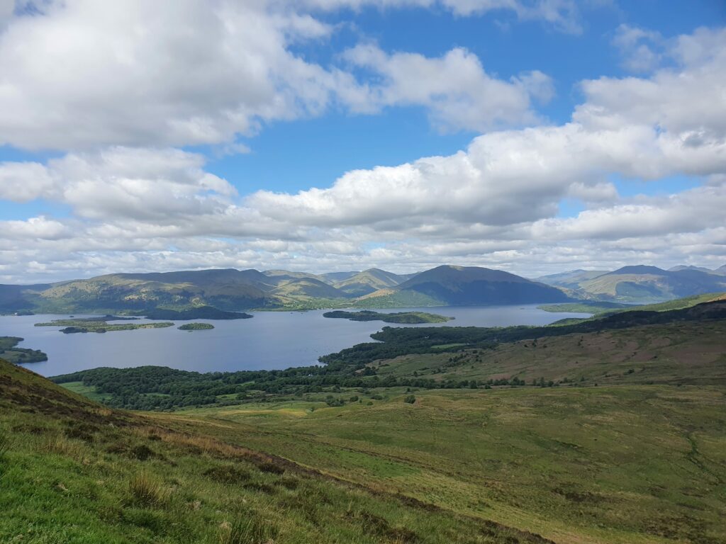 Blick vom Conic Hill über den südlichen Teil von Loch Lomond. Im Hintergrund die Ausläufer der Arrochar Alps, Im See die Inseln von loch Lomond. Der Himmel ist Blau mit weißen Wolken