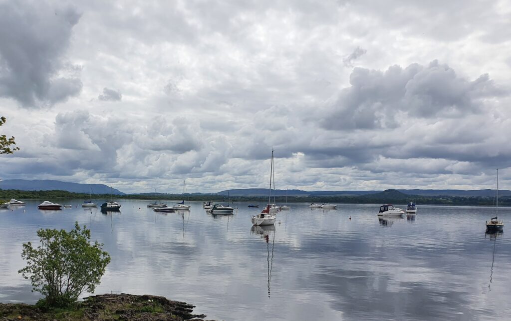 Blick über Loch Lomond. Der Himmel ist bewölkt. Im Wasser treiben Boote. 