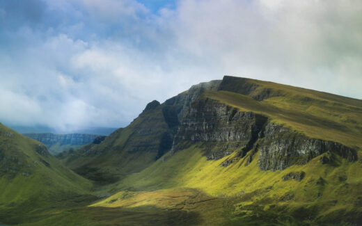 Blick ins Quiraing. Der Himmel ist blau, mit grauen Regenwolken bedeckt. Die Felswände sind dunkelgrau und darunter mit Gras bewachsen